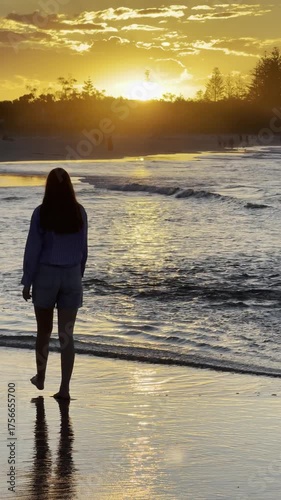 girl in elegant shirt walks in shallow water at beach admiring stunning orange sunset behind mountain ranges at popular Byron Bay Beach, New South Wales, Australia