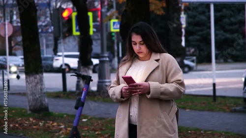 brunette woman wearing a coat standing in the street using mobile app to rent a scooter