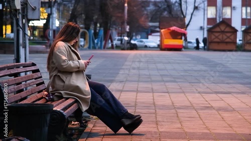 brunette woman wearing coat sitting on the bench in the street at night using smartphone