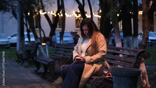 brunette woman wearing coat sitting on the bench in the street at night using smartphone