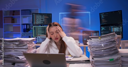Asian businesswoman working overtime at modern office desk. Piles of paperwork grow rapidly as colleagues rush past. pressure and work overload. Young woman works late at night.