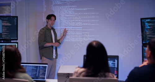 Asian male developer presenting source code to colleagues in a dark tech office. Team listening to coding explanation during software training, AI session, or cybersecurity presentation.