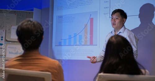 Confident Asian business professionals giving data-driven presentations to team in dark corporate office. Showing analytics charts, financial growth, and global strategy under projector light.