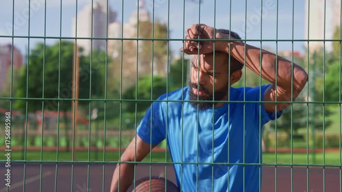 Portrait of handsome sporty fit bearded African American basketball player in deep thoughts with hand on forehead leaning on sports metal fence, looking with pensive expression outdoors.