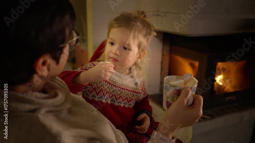 Cozy winter evening by the fireplace - grandmother and grandson share warm tea and cookies near a glowing Christmas tree. Peaceful family holiday moment filled with love, comfort, and festive lights.