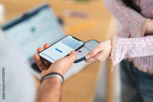 A person holds a smartphone displaying a payment interface, while another person's hand reaches out to tap their own device for a contactless transaction.