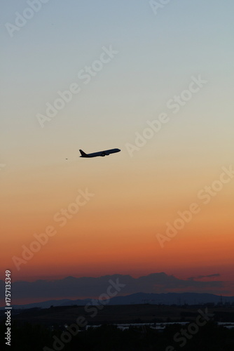 Paisaje, horizonte al atardecer en tonos degradados naranjas, con algo de nubes, se ve un avión cruzando el espacio visual, y el fondo montañoso como silueta, vertical