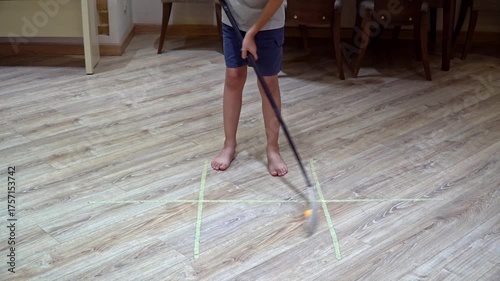 A 9-year-old boy practices hockey at home in the living room, moving the puck in a circular dribble with the stick on the floor, medium shot.