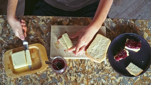 A 9-year-old boy sits at the kitchen table preparing a sandwich, cutting bread, spreading butter, adding raspberry jam with a spoon, medium closeup with selective focus and soft evening light.