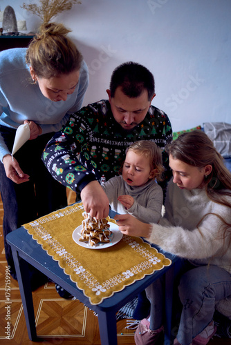 a family with daughters toddler and teenager make a Christmas tree from gingerbread cookies and decorate it with white cream and blue sugar snowflakes