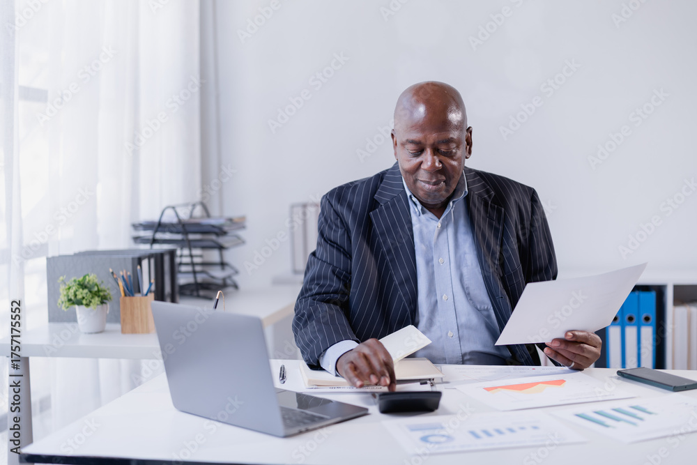 Fototapeta premium Senior African American businessman working at his office desk, reviewing financial documents and using a calculator while managing accounting and important paperwork for business analysis