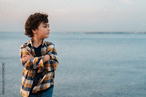Fotomural A contemplative child with crossed arms stands by the sea, gazing thoughtfully at the horizon as the sky turns pastel shades at dusk