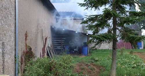 Smoke rising from building shed exterior. Fire smoke coming out of house. Building exterior with  smoke cloud. Barn on fire with thick smoke outdoors.