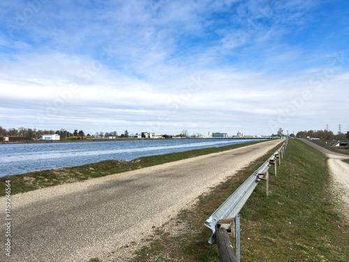 Wallpaper Mural View of the Rhine River in France with a gravel path and metal guardrail alongside waterway under a partly cloudy blue sky with industrial buildings Torontodigital.ca