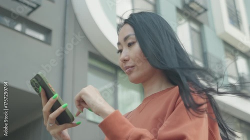 Young Asian Woman Swiping Down On Smartphone While Standing In Urban Environment