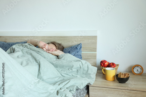 Early in the morning, a person wakes up and rubs their eyes while lying in bed. On the bedside table are an alarm clock, a yellow mug, fresh peaches, and some cookies, creating a cozy morning mood.