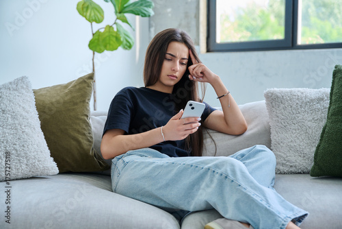 Upset sad young woman looking at smartphone sitting on couch at home
