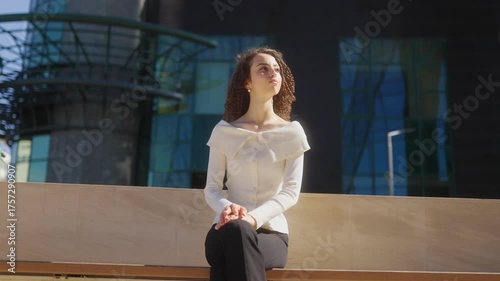 Stylish curly haired woman sitting on bench outdoors in urban sunlight
