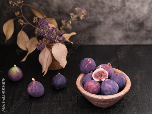 Half sliced fig in focus with whole figs nearby in a wooden bowl on dark table, rustic still life with vase decor in background