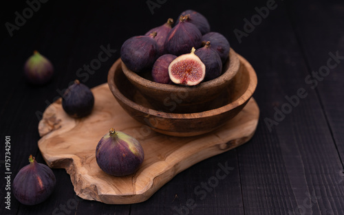 Half sliced fig in stacked bowls on wooden board with whole figs placed diagonally toward background, moody dark still life
