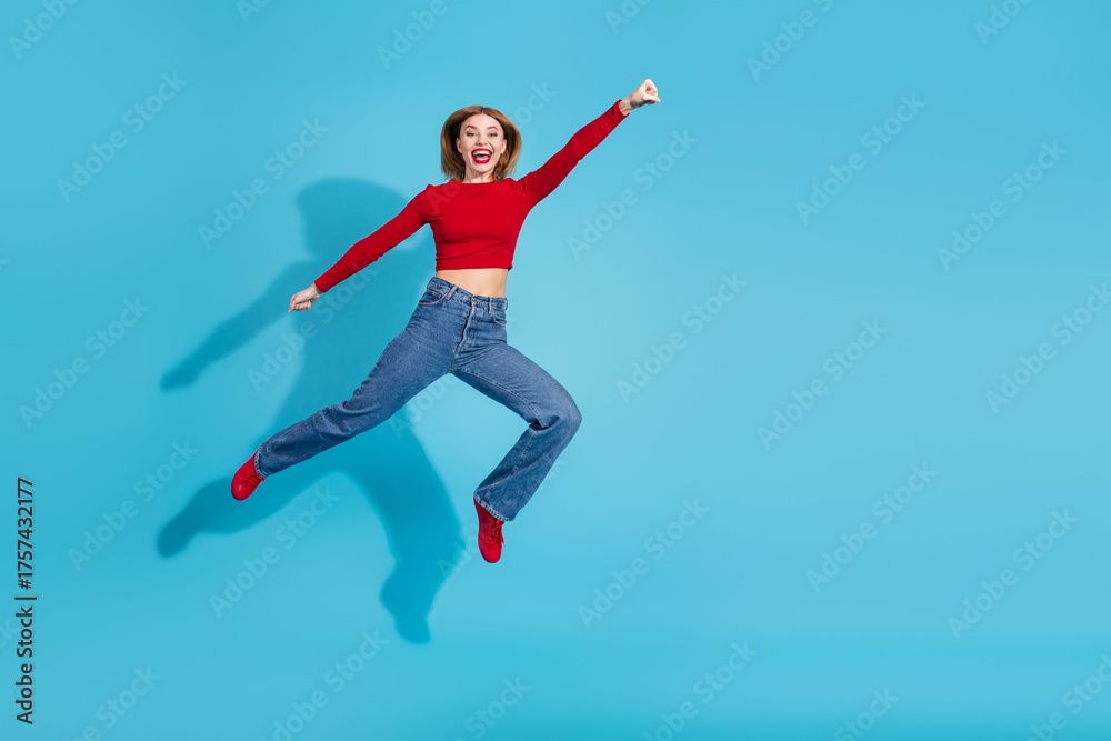 Fototapeta premium Energetic young woman in casual attire leaping joyfully against a vibrant blue background celebrating happiness