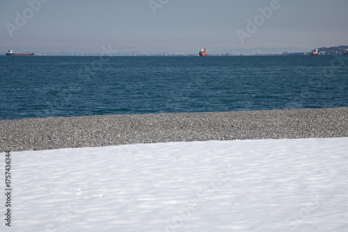 Snow covered pebble beach shoreline with cargo ships on blue sea horizon