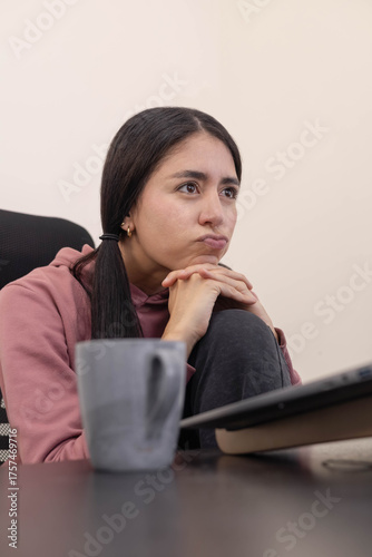 Obraz na plátně A young woman at her office desk gazes at her laptop screen with boredom, showing a lack of engagement while performing daily professional tasks in a contemporary corporate environment