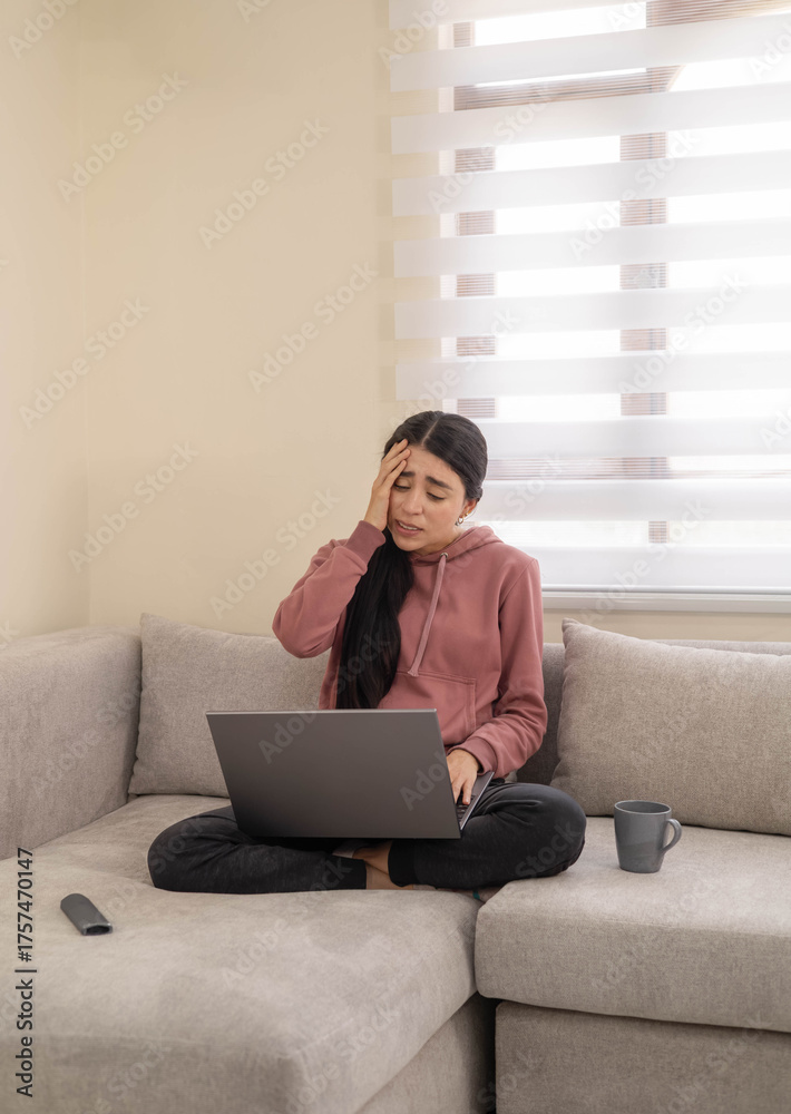 Naklejka premium A woman sitting cross-legged on a couch looks upset while viewing something on her laptop. Depicts emotional reaction, sadness, or unexpected news.