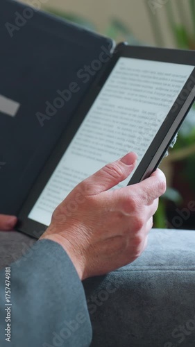 Middle-aged woman reading digital book on e-reader while relaxing on gray couch surrounded by indoor plants in cozy living room environment during peaceful afternoon moment of solitude