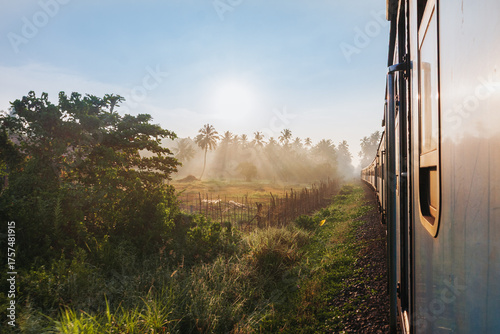 View of golden sunlight piercing through the misty landscape as seen from a train window, casting long shadows across the tracks, Kandy, Central Province, Sri Lanka.