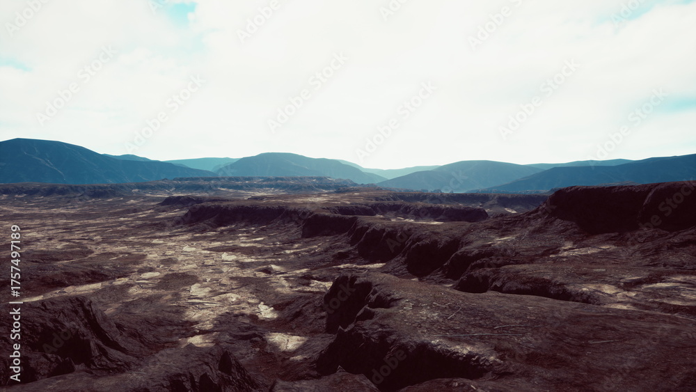 Obraz premium A vast expanse of rugged terrain stretches under a cloudy sky. The dramatic mountains rise in the background while deep valleys cut through the land, showcasing natures raw beauty.