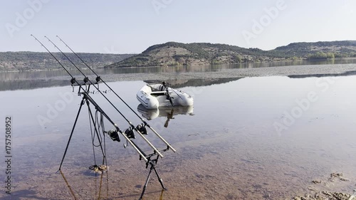 Still shot showing carp fishing rods set on bank beside a small inflatable boat by a calm lake. Peaceful freshwater fishing scene at rest.