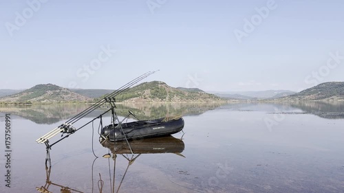 Still shot showing carp fishing rods set on bank beside a small inflatable boat by a calm lake. Peaceful freshwater fishing scene at rest.