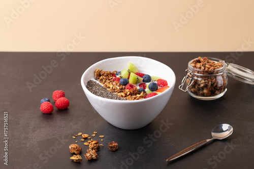 Front horizontal view of a white bowl with kefir, chia seeds, granola and fresh fruits such as kiwi, raspberries and blueberries on beige background and dark surface. Copy space. 