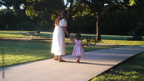 Mother and Small Toddler Daughter Holding Hands and Yellow Flowers While Walking Along Sidewalk in Green Park at Golden Hour of Sunset