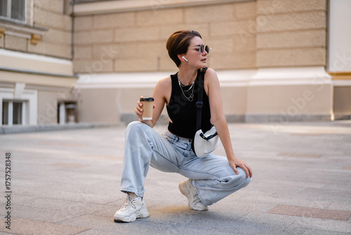 stylish woman with short haircut walking in street with coffee wearing black top, jeans and sunglasses accessories
