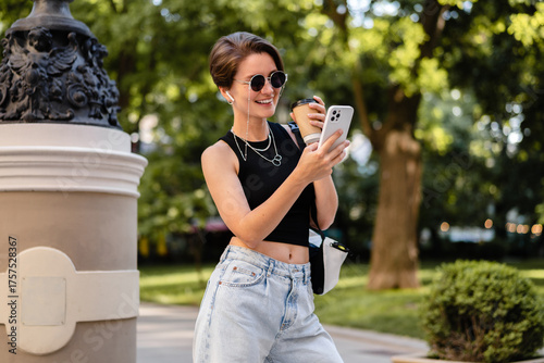 stylish woman with short haircut walking in street with coffee wearing black top, jeans and sunglasses accessories