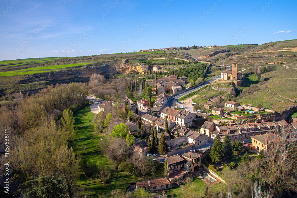 Naklejka premium A picturesque view overlooks the Segovia Templar Church of Vera Cruz in a hilly valley, surrounded by a small village of brown-roofed houses below