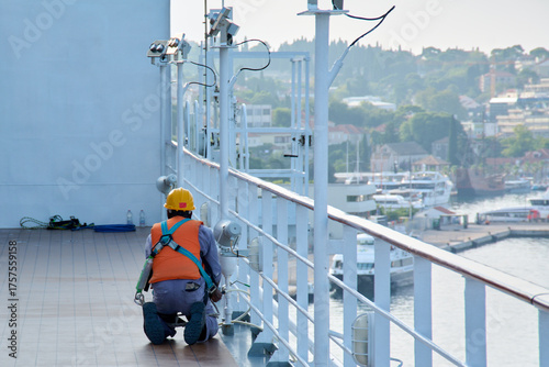 A crew member seen from behind, kneeling and wearing a safety harness, performs maintenance tasks on a vessel's deck.

