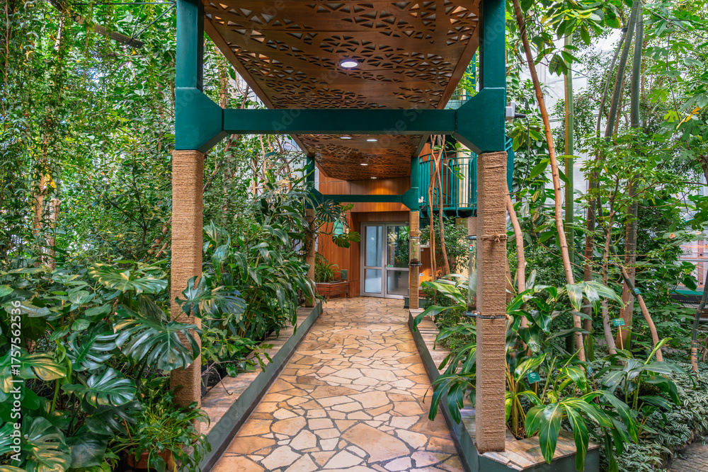 Naklejka premium Lush greenery envelops a stone pathway beneath an elevated structure, creating a serene, natural passageway in an indoor botanical setting.