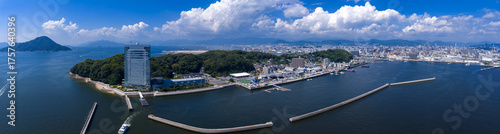 Cuadro en lienzo A wide aerial view shows Hiroshima waterfront and harbor, marinas, breakwaters, and cranes, with a conical island in the Seto Inland Sea and blue mountains under summer clouds