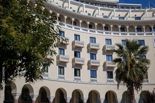 the curved, neoclassical facade of the electra palace hotel in greece, featuring arched windows, balconies, and a palm tree, framed by lush foliage