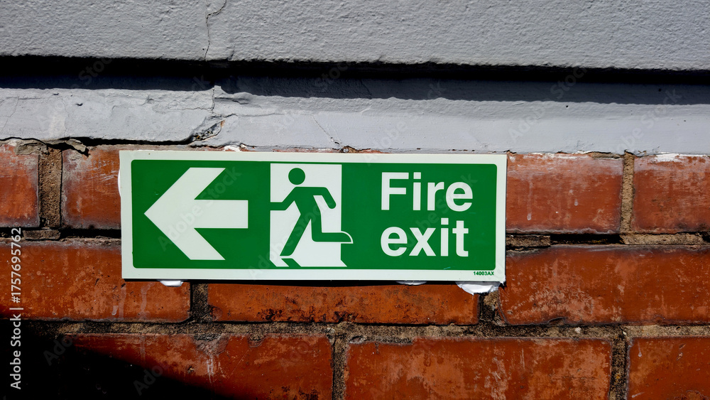Obraz premium Bright green Fire exit sign with running man pictogram and arrow pointing left on a weathered red brick wall for themes like safety, emergency, escape or public building regulations