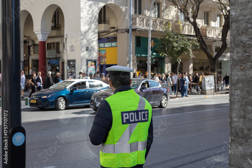 greek traffic police officer in neon vest and cap, rear view, controlling traffic on a busy european city street with cars, pedestrians, and classic arcades