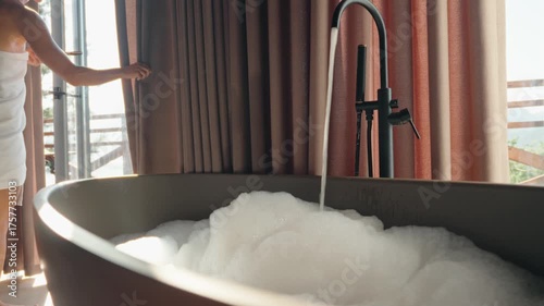 Young woman wrapped in a towel prepares a warm bubble bath in a cozy wooden country house. Morning ritual of relaxation and self-care. Natural light fills the room, creating a peaceful atmosphere