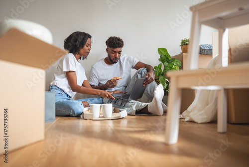 Smiling African American couple sitting on the floor among moving boxes, using a laptop together. Happy moment of love, teamwork, and excitement while planning their new home life