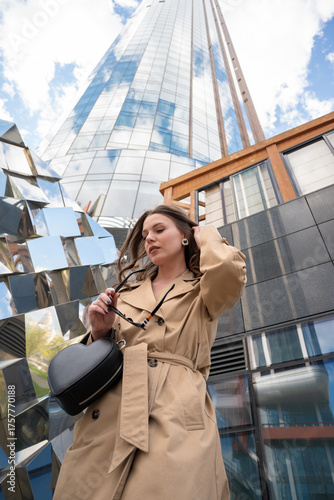 Stylish Woman in Beige Trench Coat Posing Confidently Against Reflective Glass Skyscraper