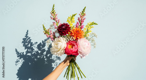 A hand holds a beautiful, elegant bouquet of fresh flowers against a bright, light blue background.