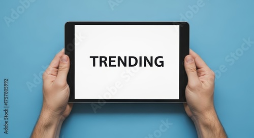 Hands holding a tablet with the word trending displayed on the screen against a blue background color