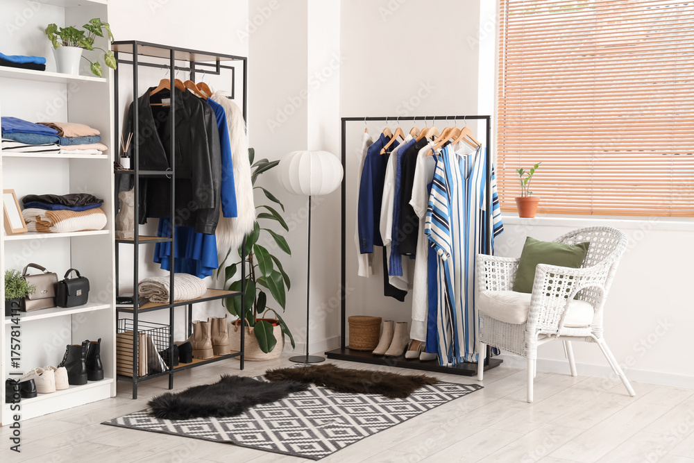 Fototapeta premium Armchair, shelving unit and racks with hanging clothes near white wall in room